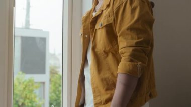 Tilt up portrait of brunet guy of 20 years old with ear piercing smiling at camera standing by window indoors wearing bright yellow jacket