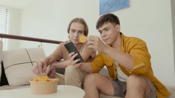 Two Buddies Eating Chips Discussing Something Smartphone Sitting Couch Home — Αρχείο Βίντεο