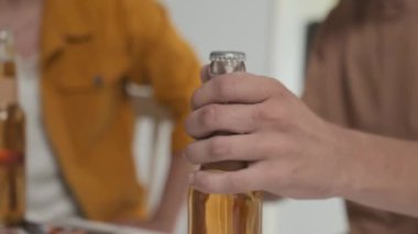 Closeup of guy opening beer bottle and toasting with male friend, hanging out together at home