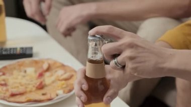 Unrecognizable guy opening glass beer bottle while hanging out with friend at home