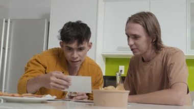 Two best mates clinking beer bottles while looking at photos together sitting by kitchen table indoors
