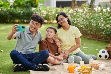 Smiling man taking selfie with his wife and little son when they are enjoying picnic in park