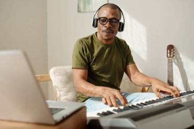 Mature man watching video on laptop when traying to learn playing synthesizer at home