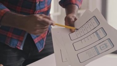 Close up shot of male mobile app developer explaining UX design sketches on paper to colleague as he taking notes at desk in office