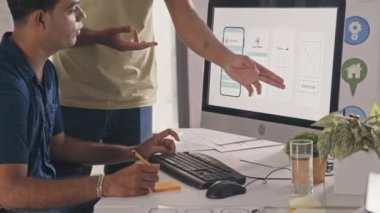 Indian UX designer discussing mobile app wireframe on computer screen with colleague while working together on project in IT company office