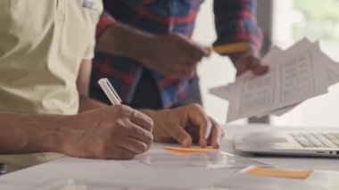 Cropped shot of two mobile app developers discussing UX project sketches on paper and laptop screen and taking notes while cooperation during workday in office