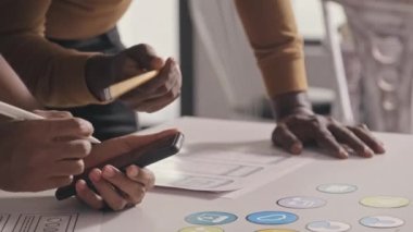 Close up shot of mobile app developers using smartphone and making notes on sketches while working together on UX project in office