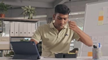Medium shot of Indian mobile app developer eating from lunch box and working on digital tablet at desk in office