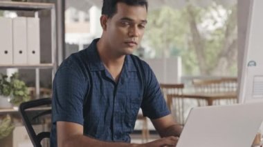 Tilt up shot of Indian programmer in casual clothes typing on laptop at office desk and then posing for camera with smile while working in IT company