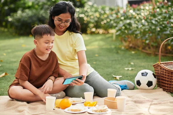 Smiling mother and son watching animated cartoon on smartphone when enjoying picnic