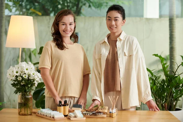 Smiling organic cosmetics shop owners standing at table with handmade soap bars and shampoes