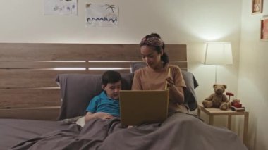Asian boy and his mother discussing photos in family album while resting together in bed in evening at home