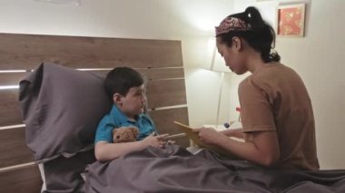Asian boy lying in bed with stuffed bear toy and listening to mother as she reading fairytale book to him before sleep