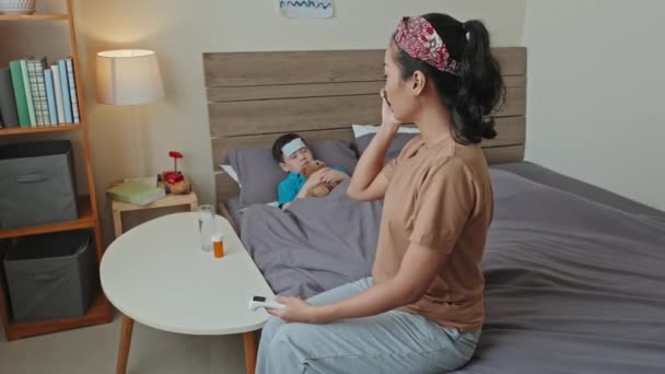 Мать Азиатка Смотрит Термометр Разговаривает Доктором Телефону Больной Сын Спит — стоковое видео