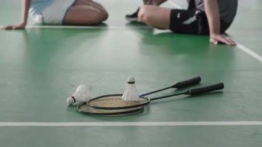 Close up shot of two rackets and shuttlecocks on floor of indoor badminton court. Unrecognizable male and female players sitting in defocused background
