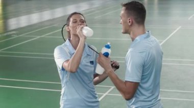 Young cheerful woman holding racket, drinking water from bottle and chatting with friend while taking break during tennis workout on indoor court