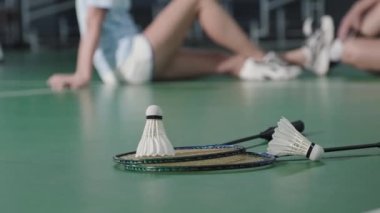 Close up selective focus shot of two rackets and shuttlecocks on floor of indoor tennis court. Unrecognizable man and woman resting in defocused background