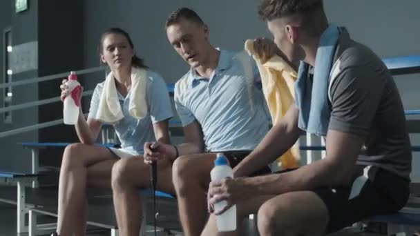 男女のテニスプレーヤーは 水とタオルでベンチに座って 屋内コートでトレーニングした後に休憩しながら話をしています — ストック動画