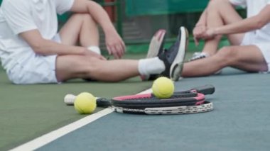 Ground level close up shot of two rackets and tennis balls on floor at outdoor court. Unrecognizable male and female players sitting in defocused background