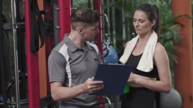 Professional male coach explaining workout plan to young sporty woman while working in gym