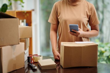 Woman checking address of client in text message when filling sticker on cardboard box for shipping order