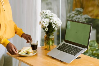 Female freelancer putting cup of coffee and plate with sandwiches on desk next to laptop