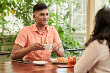 Smiling Indian man drinking coffee and talking to friends when they are sitting at cafe table