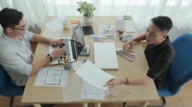 Top down shot of two young Asian men discussing blueprint and interior design plan while working together on project in office