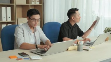 Medium shot of two young Asian architects typing on laptops and examining blueprint while working together in office