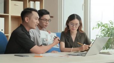 Team of young Asian coworkers discussing interior design plan via online video call on laptop while working together in office
