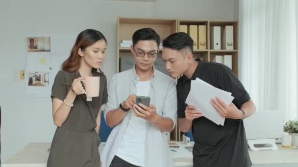 オフィスでデザインプロジェクトに取り組む一方で スマートフォンや紙で何かを議論する若いアジアの同僚のグループの中のショット — ストック動画
