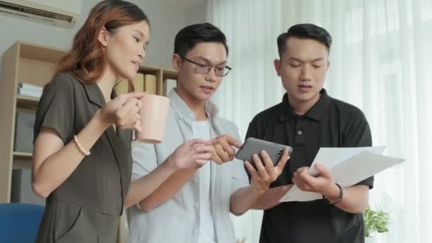 お茶を飲む若いアジアの同僚のチームの中低角度ショット オフィスでのプロジェクトに協力しながら スマートフォンや紙の上で何かを議論 — ストック動画