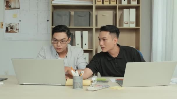 オフィスで一緒に仕事をしながら ラップトップでプロジェクトを議論する2人の若いアジアのインテリアデザイナーの中程度のショット — ストック動画