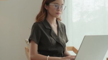 Tilt up portrait shot of young Asian woman sitting on chair at home, using laptop and then posing for camera