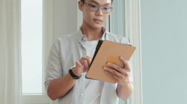 Tilt up portrait shot of young Asian man standing at home, using digital tablet and then posing for camera