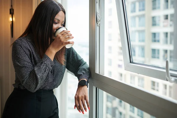 Pensive female entrepreneur drinking coffee and looking outside through window of her apartment in a skyscraper