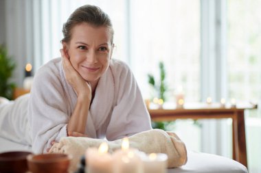 Happy middle-aged woman in soft bathrobe relaxing in spa salon with burning candles around