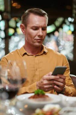Serious man sitting at restaurant table and reading message on smartphone