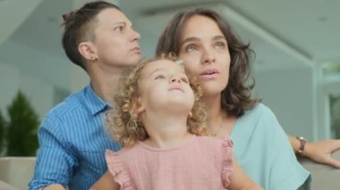 Modern Caucasian same-sex female couple with little daughter watching TV together at home, staring at screen
