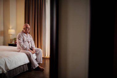 Sad depressed senior man in satin pajamas sitting on bed and staring at one point