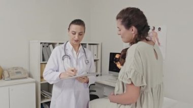 Hamile bir kadınla konuşan ve doğum öncesi kliniğinde çalışırken not alan bir kadın doktor.