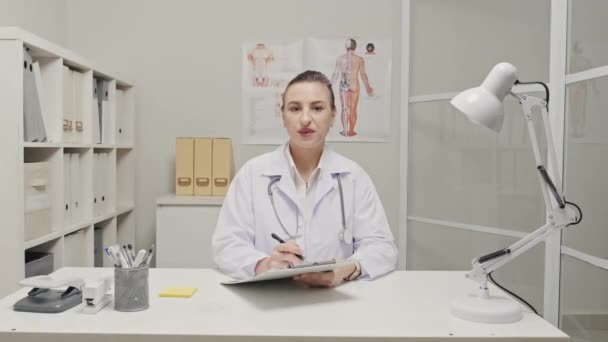 白衣を着た白人女性医師 彼女の首の上に聴診器を持つ彼女の首は医務室の机に座っています オンライン相談や撮影Vlogを与えながら クリップボードを保持し カメラで話す — ストック動画
