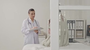 Beyaz önlüklü kadın doktorun poliklinikte doğum öncesi muayenesi sırasında elinde pano tutarken ve hamile kadını dinlerken.