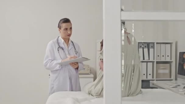 白衣姿の女性医師のアーク撮影クリップボードを保持し 診療所での出生前検査中に妊婦の話を聞く — ストック動画