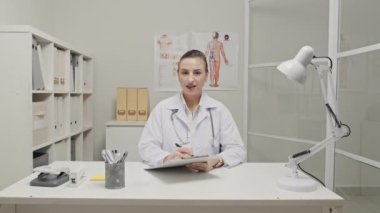 Female doctor sitting at desk in clinic, talking on camera and taking notes on clipboard while giving online consultation via video call