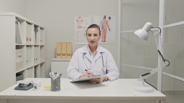 Kvinnlig Läkare Sitter Vid Skrivbordet Kliniken Prata Kameran Och Anteckna — Stockvideo