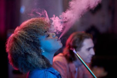Side view of curly young woman exhaling smoke wneh enjoying hookah in night clubs