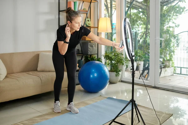 Online fitness instructor setting smartphone and ring lamp to film videos for blog at home