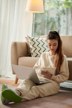 Young woman watching educational video on laptop and taking notes in planner