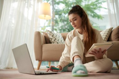 Serious young woman watching webinar on laptop and writing in notebook